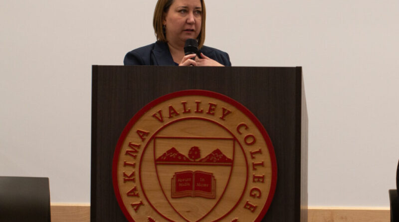 A woman stands behind a podium. She is speaking, and holding a microphone.