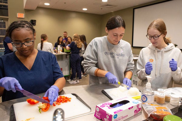 Three women with dirty purple gloves cut vegetables at a mobile kitchen table that moves into a conference room with another group of people in the background working at another prep table.